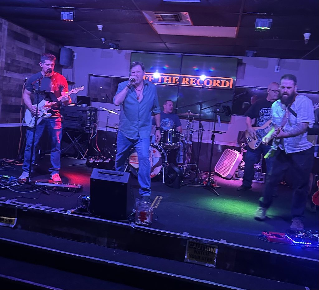 Tucson Bands and Concerts Guide (Mar 13–19, 2026) 4 Off the Record performing live on stage in Tucson with guitar, bass, drums, and vocals during a Tucson bands and concerts show.