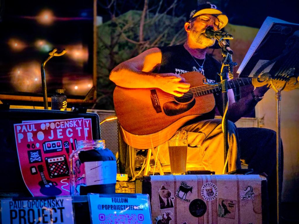 Tucson Musician Spotlight: Paul Opocensky of the PO Project 3 Paul Opocensky performing live with an acoustic guitar at Sonora Moonshine in Tucson on March 1, singing into a microphone with the Paul Opocensky Project sign beside him.