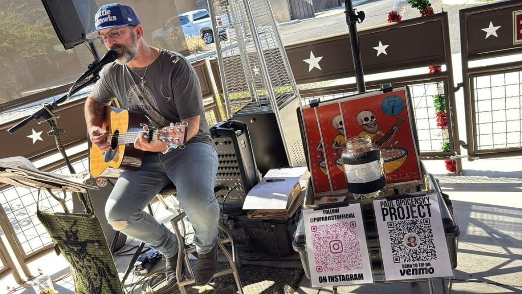 Paul Opocensky performing live with acoustic guitar at DiBella’s Brunch N Booze in Tucson in December 2025 with the Paul Opocensky Project tip jar and QR codes for fans.