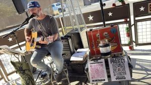 Paul Opocensky performing live with acoustic guitar at DiBella’s Brunch N Booze in Tucson in December 2025 with the Paul Opocensky Project tip jar and QR codes for fans.
