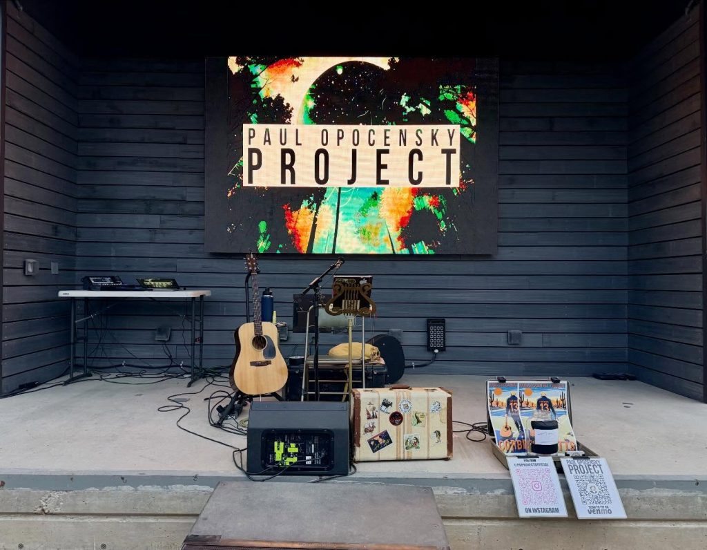 Tucson Musician Spotlight: Paul Opocensky of the PO Project 4 Stage setup for Paul Opocensky Project at Corbett’s in Tucson in 2026, featuring acoustic guitar, microphone stand, suitcase tip jar, and digital screen displaying Paul Opocensky Project.