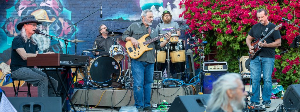 Legion of Mario performing live outdoors in Tucson, Arizona in 2024, featuring band members playing keyboard, drums, bass, percussion, and guitar in front of colorful murals and blooming bougainvillea.