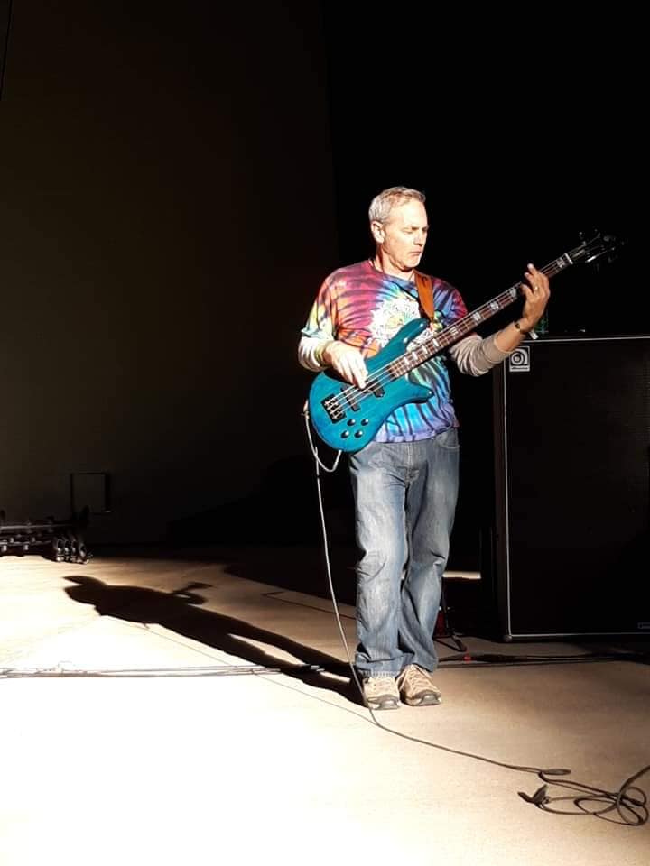 Mark Ryan performing live on bass guitar under stage lighting, wearing a colorful tie-dye shirt during an indoor concert performance in Tucson, Arizona.