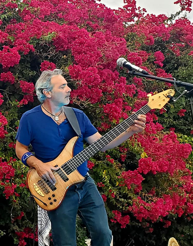 Mark Ryan performing on bass guitar at The Hut on 4th Avenue in Tucson, Arizona, standing in front of vibrant pink bougainvillea during an outdoor live music performance.