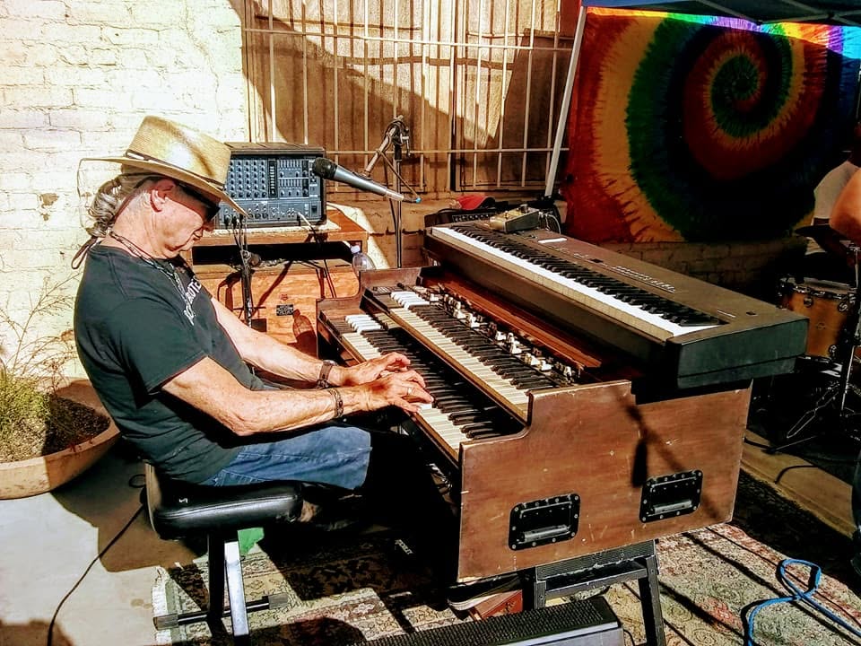 Tucson Musician Spotlight: Mark Holladay 3 "Doc" Mark Holladay performs on a Hammond organ during an outdoor Tucson music event in 2018. Wearing a hat and sunglasses, he sits at the keyboard immersed in his performance, with audio equipment, colorful tie-dye artwork, and fellow musicians visible in the background. Photo by Jack Straw.
