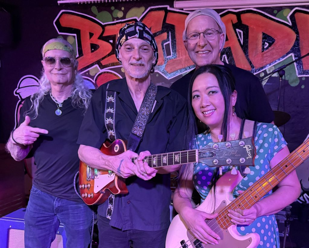 Tucson Musician Spotlight: Mark Holladay 4 Members of ORGANic Funk Alliance pose together on stage in Tucson in 2026. "Doc" Mark Holladay stands behind the group, smiling beside guitarist Eric Garcia, bassist Koko Matsumoto, and another band member. The musicians hold their instruments in front of a colorful "Big Bad Wolf" mural backdrop at a local live music venue.