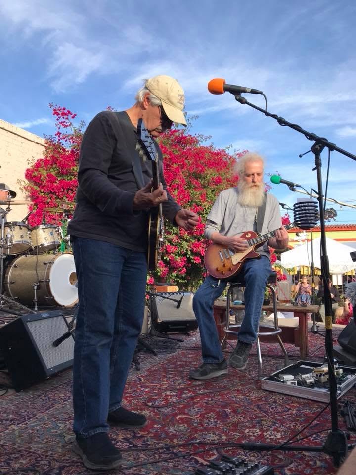 Frank Filipo performs with Randy Clamons and Top Dead Center at The Hut in Tucson in 2017. Frank stands at the front of the stage playing guitar while Randy, seated with his electric guitar, performs beside him. Bright bougainvillea flowers and blue skies provide a colorful backdrop to the outdoor live music performance.