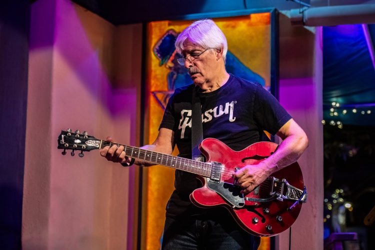 Frank Filipo performs live with Brian Fate in 2025, playing a red semi-hollow Gibson electric guitar under colorful stage lighting. Wearing a black Gibson t-shirt, Frank focuses on his performance as purple and gold lights illuminate the stage behind him. Photo by Brian Fate.