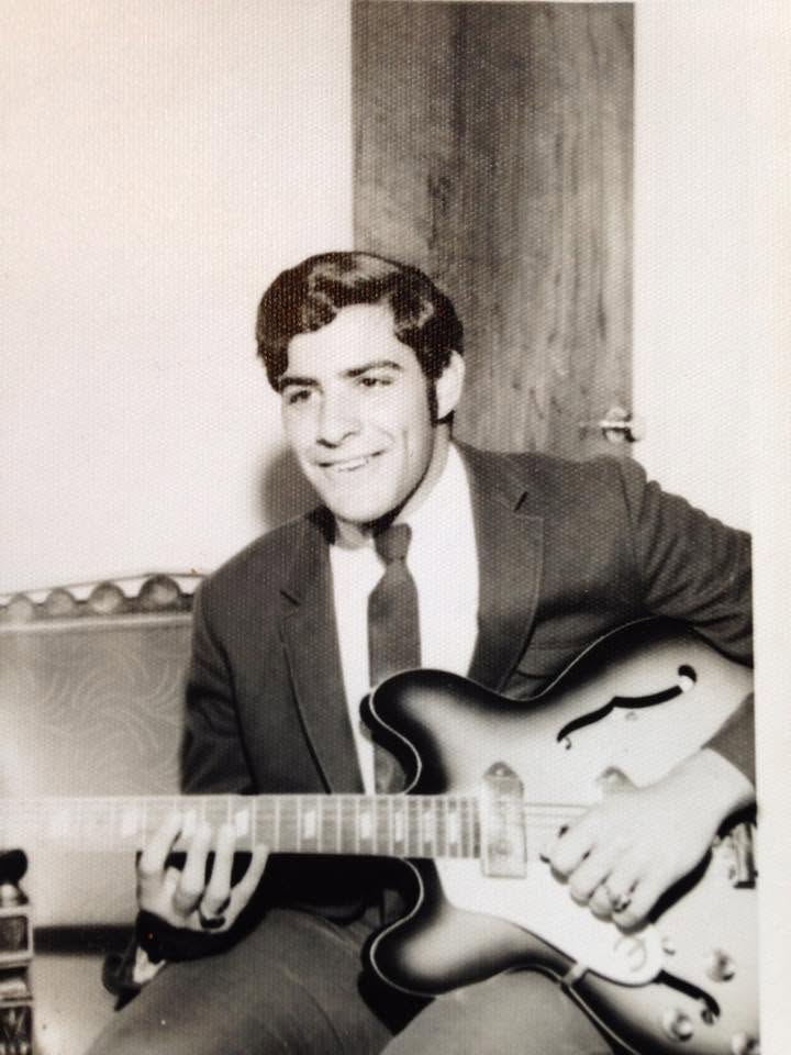 Black-and-white vintage photo of a young Frank Filipo seated with a hollow-body electric guitar. Dressed in a suit and tie, he smiles toward the camera while holding the guitar, capturing an early moment in his lifelong musical journey.