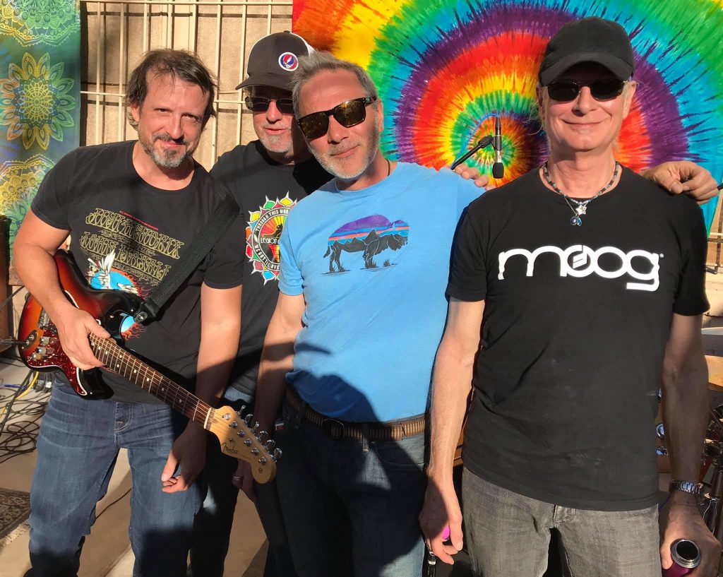 Tucson Musician Spotlight: Mark Holladay 5 Members of Legion of Mario pose together outdoors before a performance in Tucson in 2019. The band stands in front of a colorful tie-dye backdrop, with guitarist Frank Filipo holding a Fender guitar and keyboardist "Doc" Mark Holladay wearing a black Moog shirt and sunglasses. Photo by Erika Tenney.