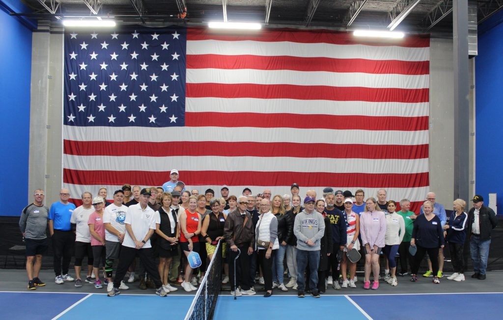 Group photo of 100-year-old Navy veteran Bill Kruschel with family members and USA 4 Veterans Foundation pickleball supporters posed in front of a large American flag at an indoor pickleball court.