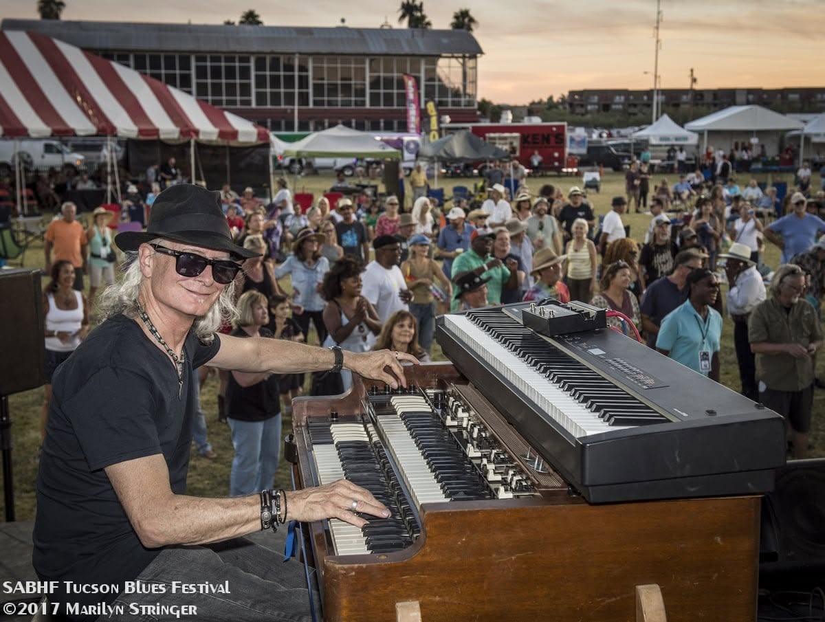 Mark "Doc" Holladay performs on his Hammond organ during the Tucson Blues Festival in 2021. Seated on stage in a black hat and sunglasses, he smiles while playing to a large outdoor crowd gathered in front of the festival stage at sunset. Photo by Marilyn Stringer.